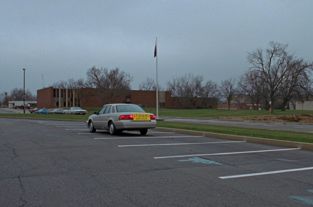 A plain sedan with a yellow student driver sign parked in an almost empty municipal lot in front of a low brick building on an overcast day.