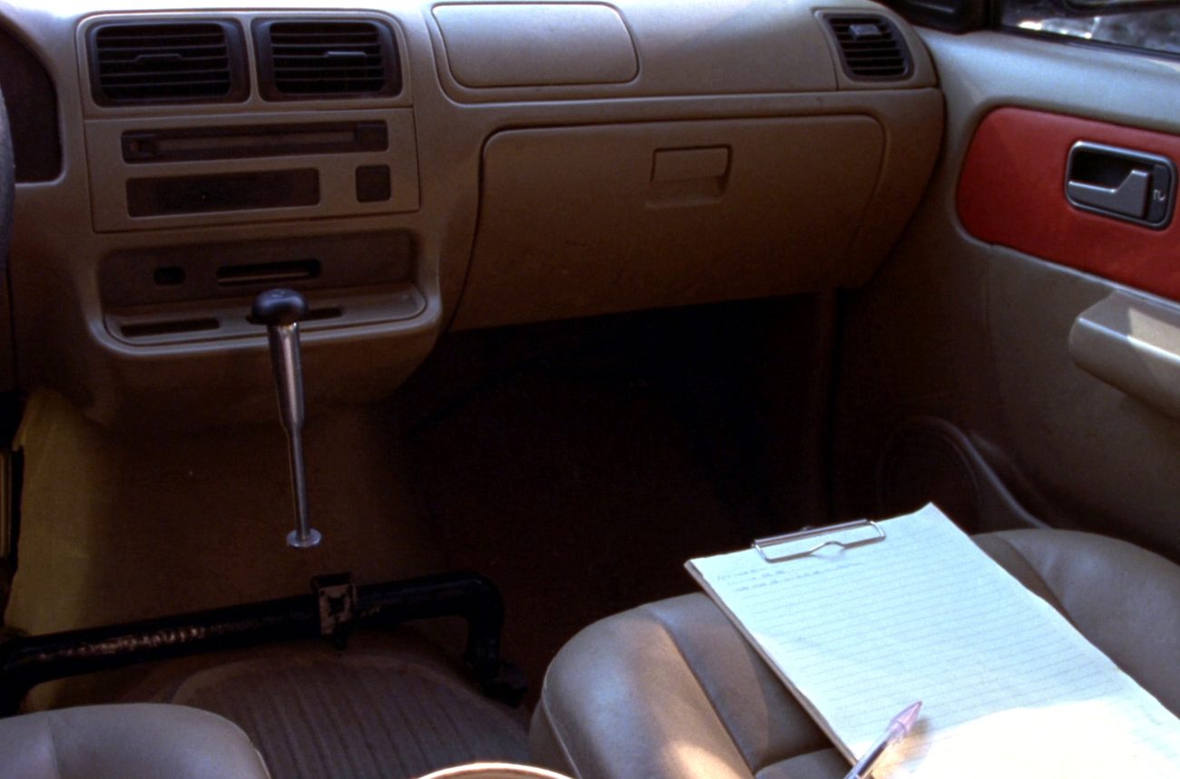 The passenger side of a training car interior, a second brake bracket fitted across the floor and a clipboard with a blank form resting on the seat.
