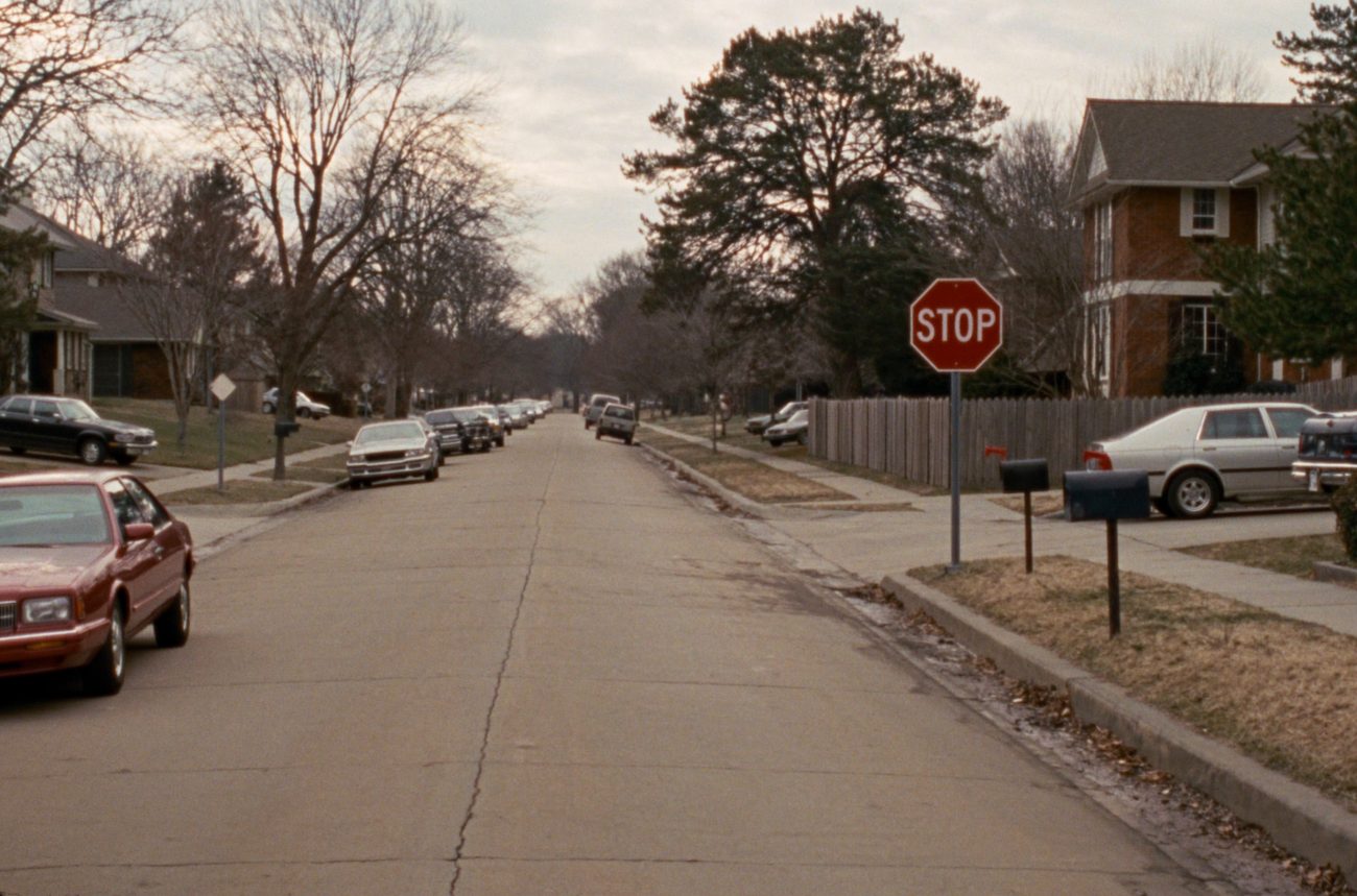 A quiet residential street on a grey winter afternoon, cars parked along both kerbs, mailboxes on the verge and a stop sign at the corner.
