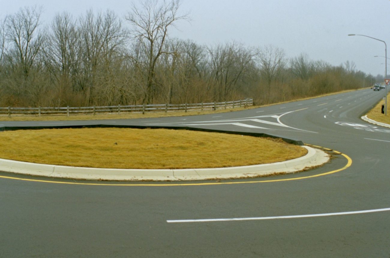 A single lane suburban roundabout photographed from a rise in flat overcast light, with painted yield lines and a grassed centre island.