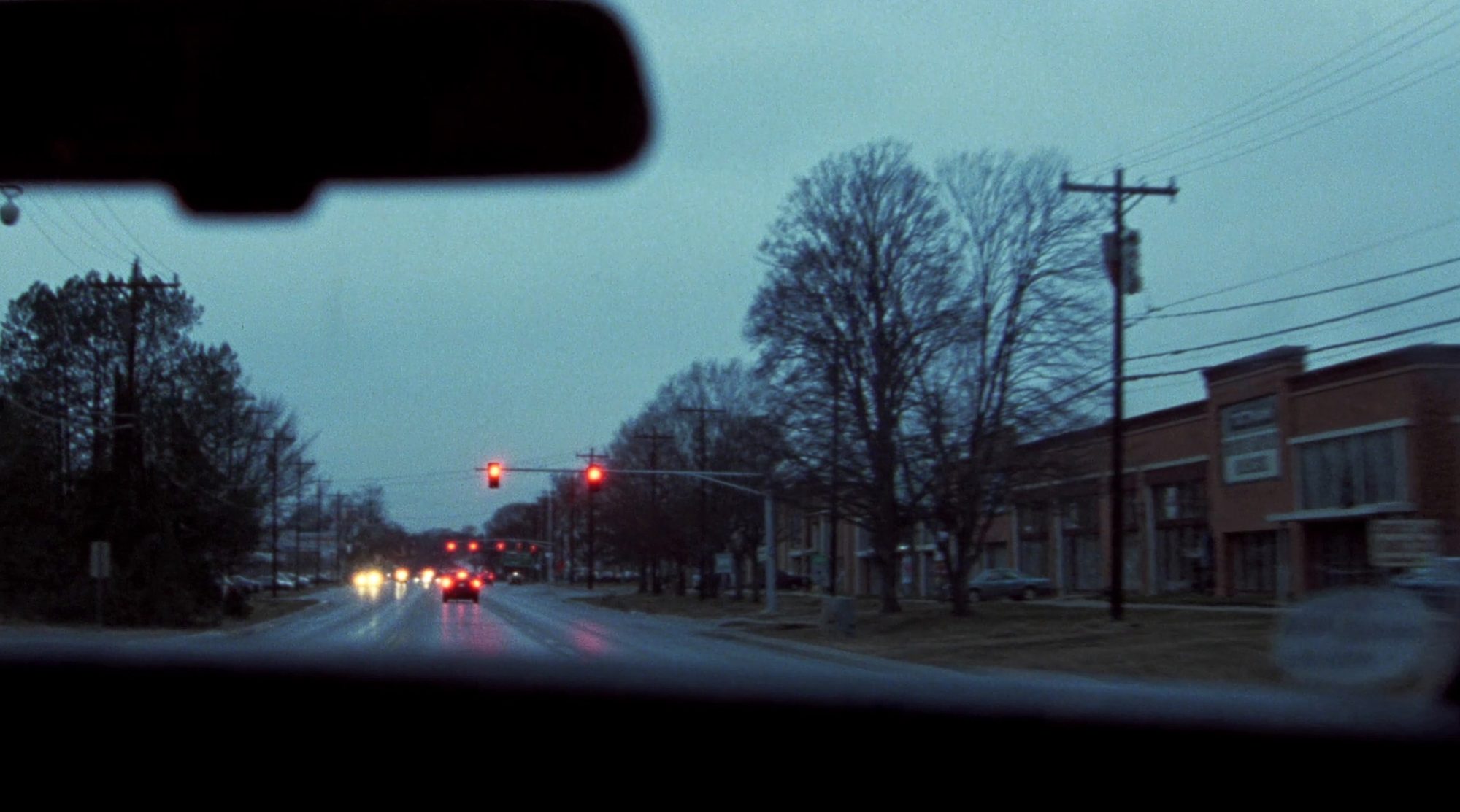 View through a car windshield at dusk on a wet four-lane suburban road, tail lights ahead and a traffic signal overhead.